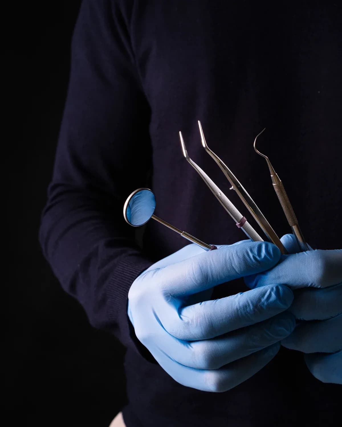 Mãos com luva segurando espelho, pinça e sonda odontológicos sobre fundo escuro